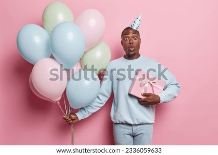 Surprised dark skinned man wears party hat blue jumper and jeans poses with birthday present and bunch of colorful balloons going on party to congratulate friend isolated over pink background