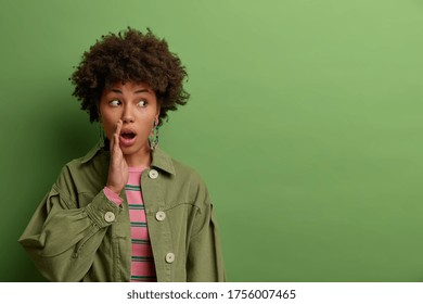 Surprised dark skinned millennial woman gossips and tells secret, keeps mouth opened, looks aside, doesnt want someone hear her story, wears fashionable clothing, isolated on green studio background - Shutterstock ID 1756007465
