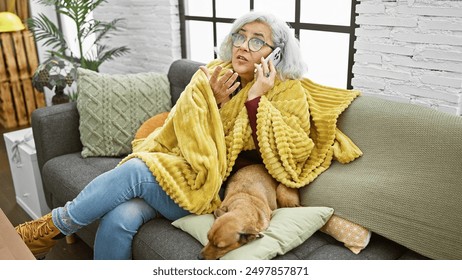 A surprised elderly woman in glasses talks on a phone, cozy in a yellow blanket with her dog beside her in a modern living room. - Powered by Shutterstock