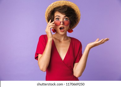 Surprised Elegant Woman In Straw Hat Take Off Sunglasses While Looking At The Camera Over Violet Background