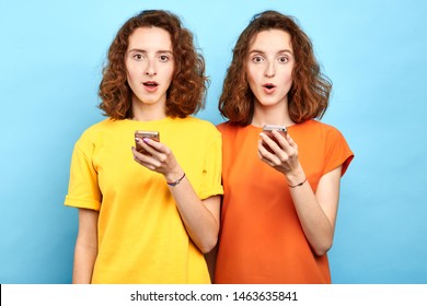surprised emotional twins with wide open mouth and bugged eyes holding smart phones and looking at the camera. sale, business, free message, double discounts
