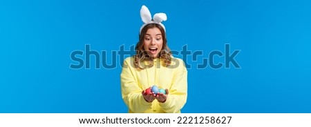 Surprised emotive cute blond caucasian girl playing holiday game, found Easter eggs look amused, wearing rabbit ears as celebrating religious holiday on sunday, blue background.