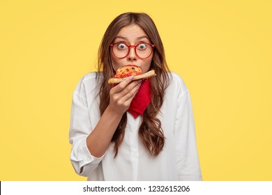 Surprised European fashionable woman has slice of pizza, looks at camera, dressed in oversized shirt, surprised with very nice taste, isolated over yellow background. People and fast food concept