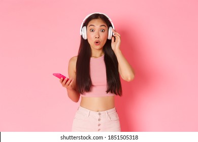 Surprised And Excited Asian Cute Girl Stare At Camera Wondered While Listening Interesting Podcast Or Friends Playlist, Checking Out New Song In Headphones, Holding Mobile Phone, Pink Wall