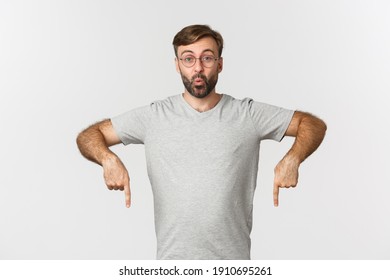 Surprised and excited bearded man, pointing fingers down, showing logo, wearing gray t-shirt, wearing gray t-shirt, standing over white background