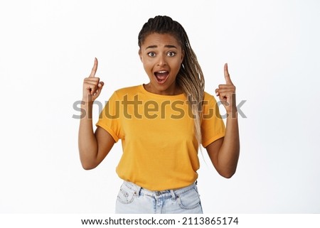 Surprised and excited Black woman pointing fingers up, gasping in awe, amazed by price sale, standing in yellow t-shirt over white background Foto stock © 