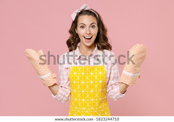 Surprised excited cheerful young brunette woman\
housewife 20s wearing yellow apron gloves potholders doing\
housework isolated on pastel pink colour background studio\
portrait. Housekeeping\
concept