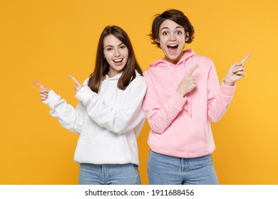 Surprised excited funny two young women friends 20s wearing casual white pink hoodies pointing index fingers aside up on mock up copy space isolated on bright yellow color background studio portrait
