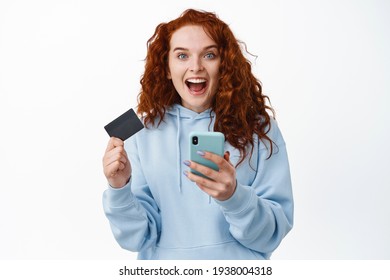 Surprised And Excited Ginger Girl With Curly Hair, Holding Mobile Phone And Plastic Credit Card, Staring Amazed At Camera, Winning Money, Checking Bank Account Balance, White Background