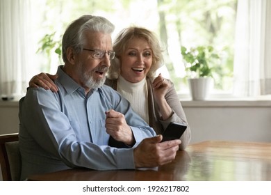 Surprised Excited OAP Family Couple Getting Amazing Good News From Video Call. Happy Middle Aged Pensioners Using Mobile Phone Together At Home, Talking To Relations, Looking At Screen And Gasping