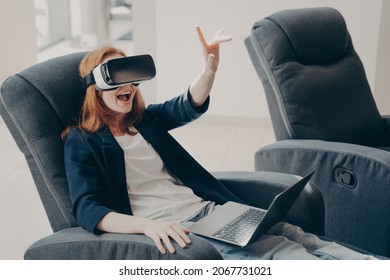 Surprised Excited Red Haired Woman In VR Headset Goggles And Laptop, Sitting In Soft Big Armchair In Office, Trying To Touch 3D Virtual Reality Objects, Pointing With Index Finger Up In Air