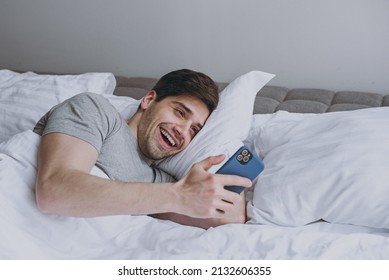 Surprised Excited Young Man In Pajamas Grey T-shirt Lying In Bed Sleep Read Message On Mobile Cell Phone Chatting Online Rest Relax At Home Indoors Bedroom. Good Mood Night Morning Bedtime Concept.