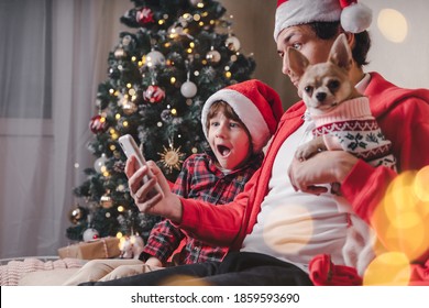 Surprised Father With Kid And Puppy Dog In Santa Hats Holding Cell Phone On Christmas Holidays, Sitting On A Couch In The Living Room With Christmas Tree At Home.