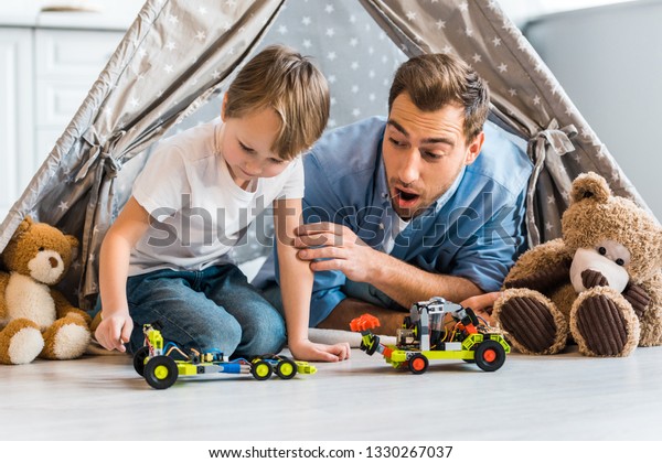 surprised father looking at preschooler son\
playing with toy car under wigwam at\
home