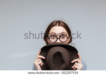 Surprised Female nerd in eyeglasses which looking behind the hat over gray background