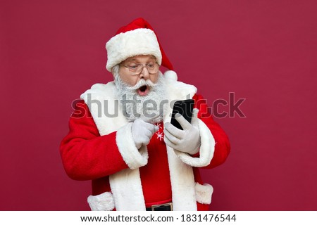 Surprised funny amazed old bearded Santa Claus wearing costume holding cell phone using mobile app on smartphone shocked by Christmas promotion, xmas applications ads isolated on red background.