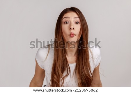 Surprised funny woman with dark hair making fish face grimace with pout lips and looking with confused comical expression, wearing white T-shirt. Indoor studio shot isolated on gray background.