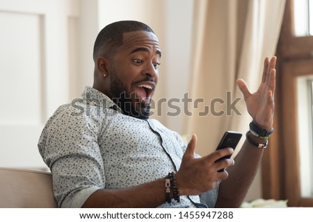 Surprised happy african man holding phone looking at cellphone read good news in sms sit on sofa, amazed black guy winner excited by mobile app win scream with joy celebrate victory success at home