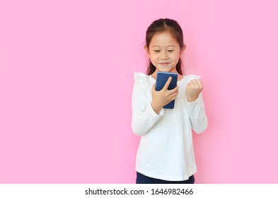 Surprised Happy Asian Little Child Girl Watching Her Smartphone And Expression Hand Fight Sign Over Pink Background With Copy Space. Kid Smiling And Enjoy While Playing Smart Phone.