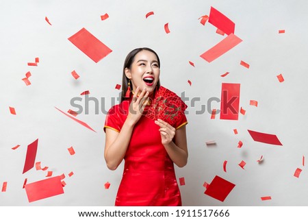 Surprised happy Asian woman in traditional oriental costume holding red envelopes or Ang Pao in gray background with confetti, Chinese text means great luck great profit