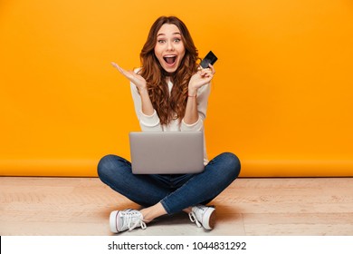 Surprised happy brunette woman in sweater sitting on the floor with laptop computer while holding credit card and looking at the camera over yellow background