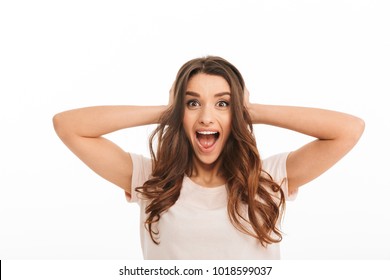 Surprised happy brunette woman in t-shirt screaming and covering her ears while looking at the camera over white background