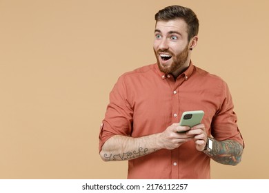 Surprised happy tattooed young brunet man 20s he wears orange shirt hold in hand use mobile cell phone look aside isolated on plain pastel light beige background studio portrait. Tattoo translate fun