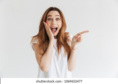 Surprised happy woman in t-shirt holding her cheek and pointing away while looking at the camera over grey background