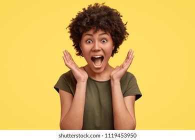 Surprised happy young dark skinned female stands in big shopping mall, glad to see big discounts on clothes, expresses shock, keeps hands near head, jaw dropped, going to spend much money today