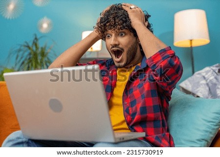 Surprised indian man guy using laptop computer, receive mail good news message, shocked by sudden victory celebrate lottery jackpot win purchases online shopping at home apartment on sofa. Lifestyles