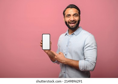 Surprised Indian Man Holds Smartphone In A Hand And Points Finger At Empty Phone Screen, Astonishment Hispanic Guy Presenting Best Deal, Advertising New Mobile App, Isolated On Pink