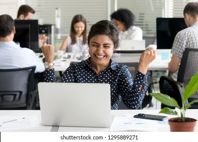 Surprised indian worker sitting at desk in coworking space with colleagues looking at notebook screen feels excited and amazed. Female received unexpected great opportunity promotion or getting reward
