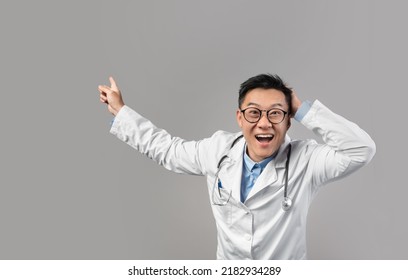 Surprised Inspired Mature Chinese Man Doctor In White Coat, Glasses With Open Mouth Pointing Finger At Free Space Isolated On Gray Background. Human Emotions, Health Care, Huge Medical Ad And Offer