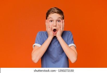 Surprised Little Boy. Shocked Kid Keeping Mouth Open And Touching Face, Orange Studio Background