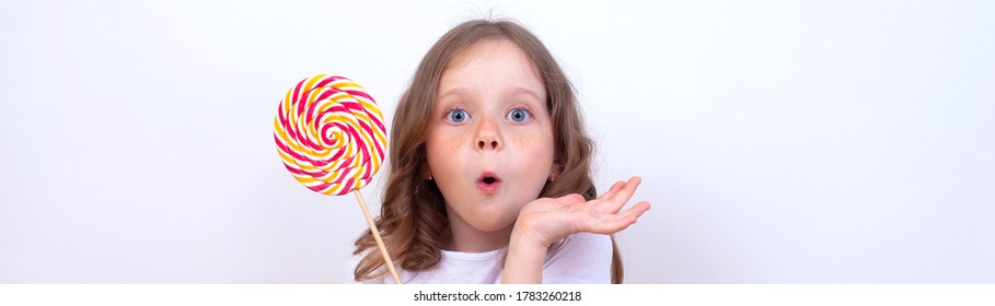 A surprised little girl holds a large Lollipop in her hand. Beautiful baby face on a large banner background with copy space. extra wide