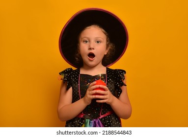 Surprised Little Girl Wearing Halloween Witch Costume Or Wizard Costume And Holding Red Lighted Candle In Hand On A Yellow Background.

Cute Amazed Kid With Open Mouth Looking At Camera.