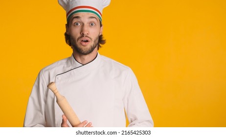 Surprised Male Chef In White Uniform And Cap Holding Wooden Rolling Pin Looking Amazed At Camera Over Orange Background. Young Chef Not Ready For Making A Cake