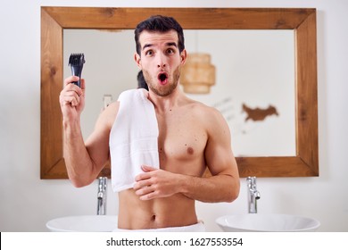 Surprised Man With Electric Razor In Hand With Towel On His Shoulder Stands Near Mirror In Bath