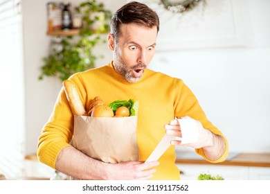 Surprised man looking at store receipt after shopping, holding a paper bag with healthy food. Guy in the kitchen. Real people expression. Inflation concept.