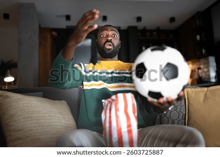 A surprised man reacts dramatically while watching a soccer match on TV at home. He sits on a couch with a bowl of popcorn as a soccer ball seemingly comes toward him, adding excitement to the moment.