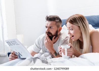 Surprised Man And Young Woman With Cup Lying In Bed And Reading Travel Newspaper