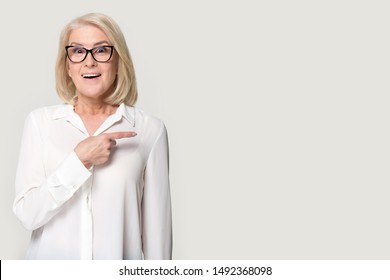 Surprised Mature Lady Wearing Glasses Blouse Isolated On Grey Studio Background Point At Blank Copy Space Aside, Senior Woman In Spectacles Feel Shocked By Amazing Good Promotion, Sale Offer Or Deal