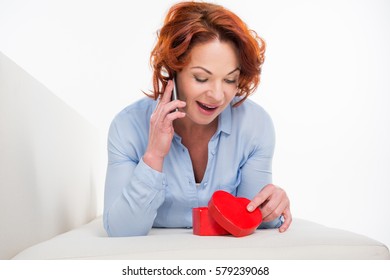 Surprised Mature Woman Talking On Smartphone While Opening Red Heart Shaped Gift Box Isolated On White