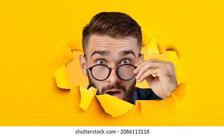 Surprised millennial caucasian guy takes off his glasses and looks in camera through hole in yellow paper, panorama, collage, close up. Shocking offer and advertisement, sale and facial expressions