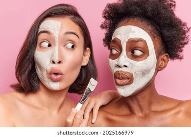 Surprised mixed race friendly women apply nourishing face masks with cosmetic brush look shocked at each other stand shirtless against pink background. Skin care beauty and wellness concept.