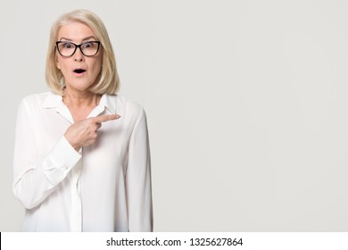 Surprised Old Mature Businesswoman Pointing Finger At Copy Space Looking At Camera, Amazed Senior Middle Aged Woman In Glasses Advertising Product Service Isolated On White Grey Studio Background