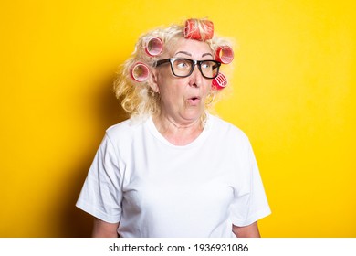 Surprised Old Woman With Curlers In Glasses, In A White T-shirt On A Yellow Background.