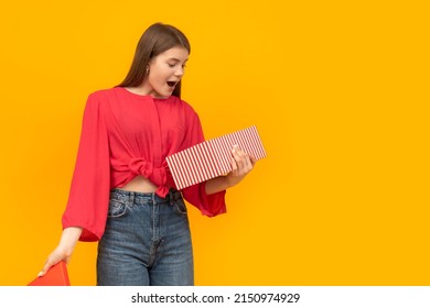 Surprised And Overwhelmed Young Girl With Gift Box In Her Hands. Yellow Background. Unexpected Gift