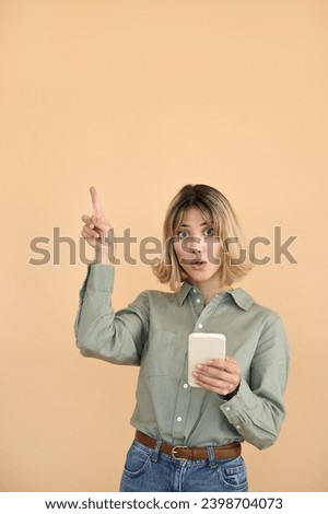 Surprised pretty gen z blonde young woman with smartphone, amazed 20s european customer girl with short blond hair wow expression pointing up using mobile cell phone on beige background. Copy space.
