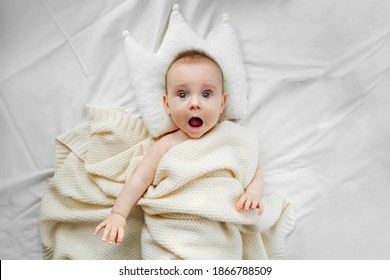 Surprised or scared baby lying in bed with princess pillow. Infant baby wrapped in warm knitted blanket lying on white cozy bed. View from above